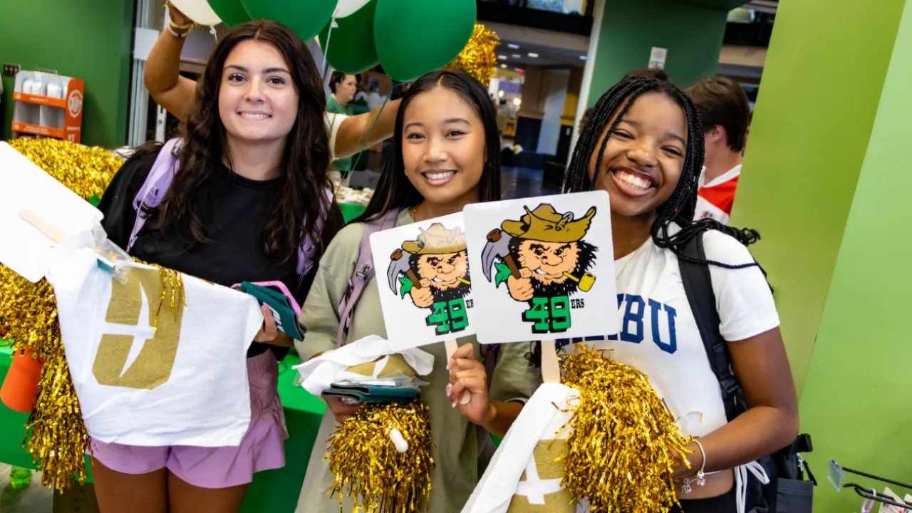 three students at a Gold Rush event. They are holding swag and pom poms while surrounded by Charlotte color decorations. 