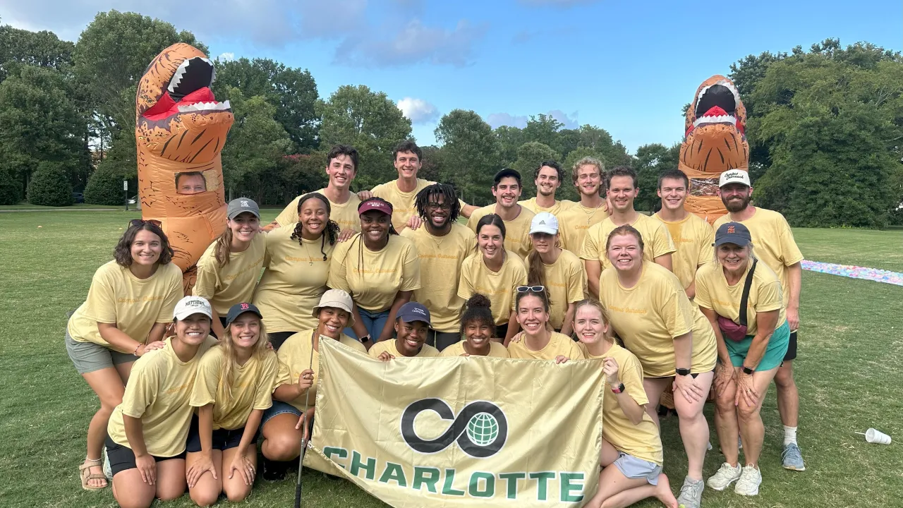 Group of students in a tableau holding a banner that has the Campus Outreach logo and Charlotte underneath. They are wearing gold shirts and there are two people in blow up dinosaur costumes flanking the students.