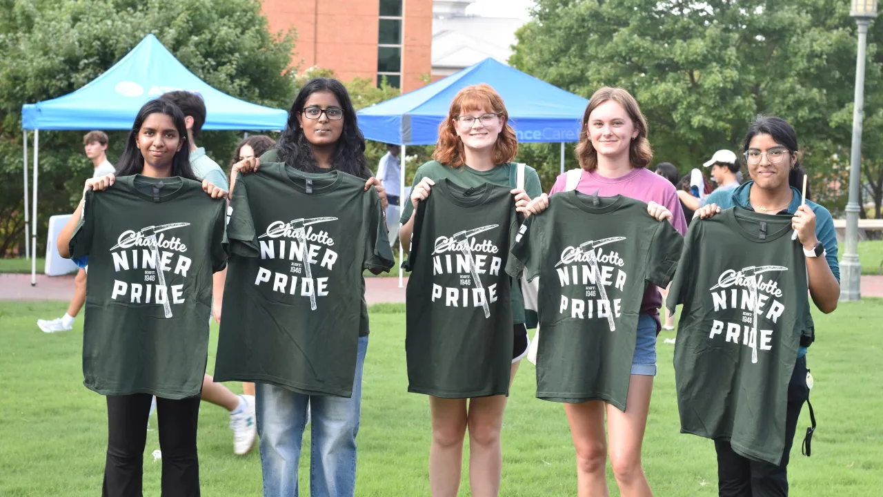 5 students holding up the 2025 - 2026 Niner Pride tshirts outside during New Student Welcome. 