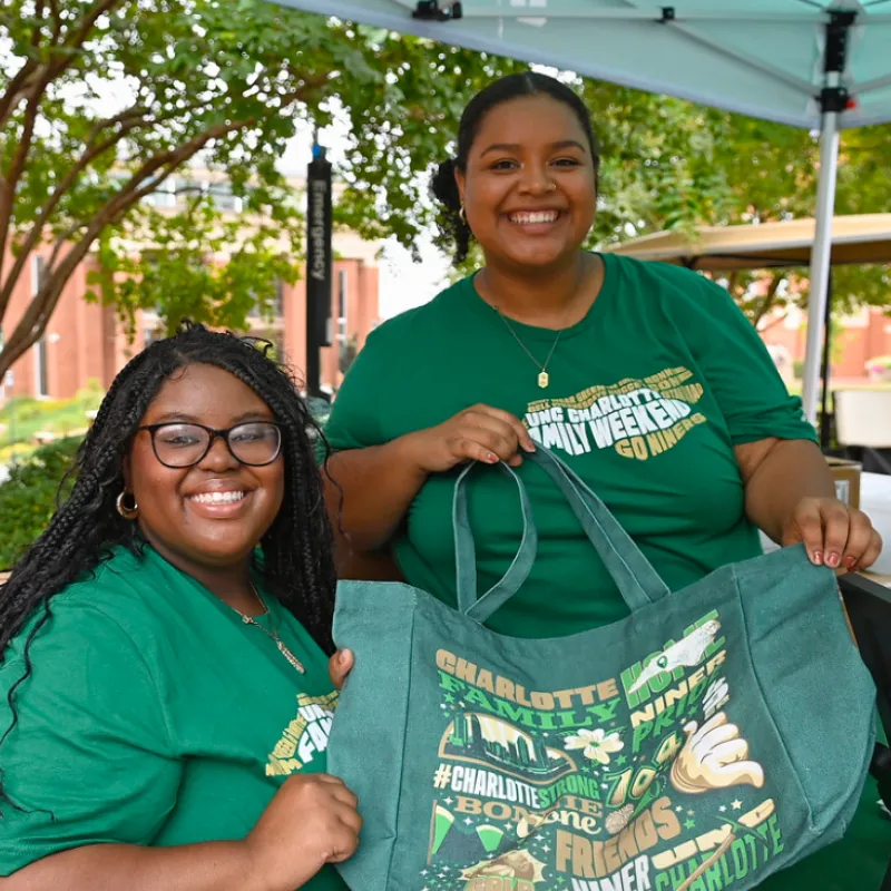 Two student leaders holding a Family tote that is green and has charlotte symbols all over it. 